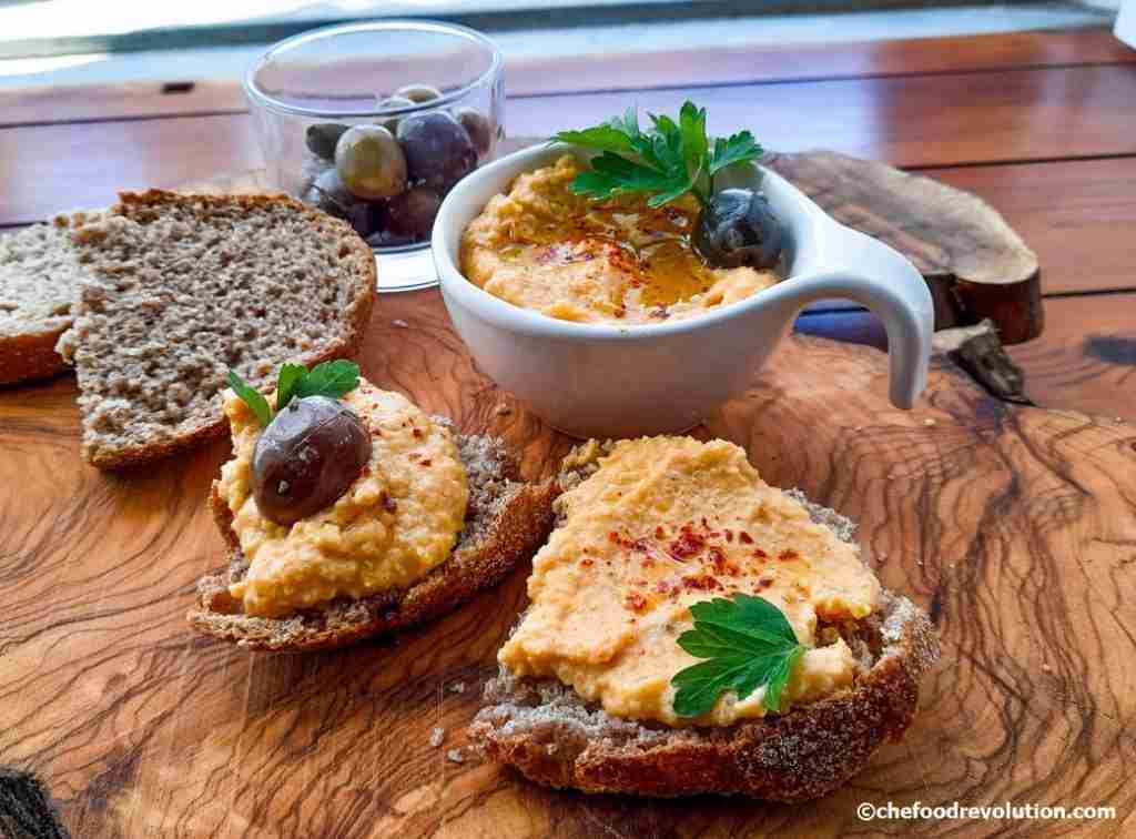 elegante composizione con ciotolina di hummus di zucca, fette di pane, olive, il tutto su un tagliere di legno di ulivo